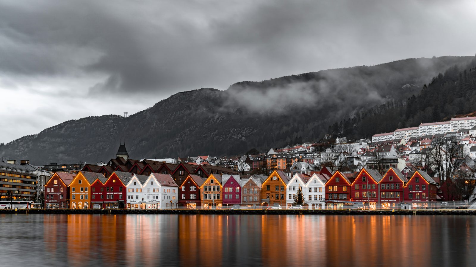 Bryggen-husene opplyst i kveldsmørket, speilet i Vågen, med tåke over byfjellet bak