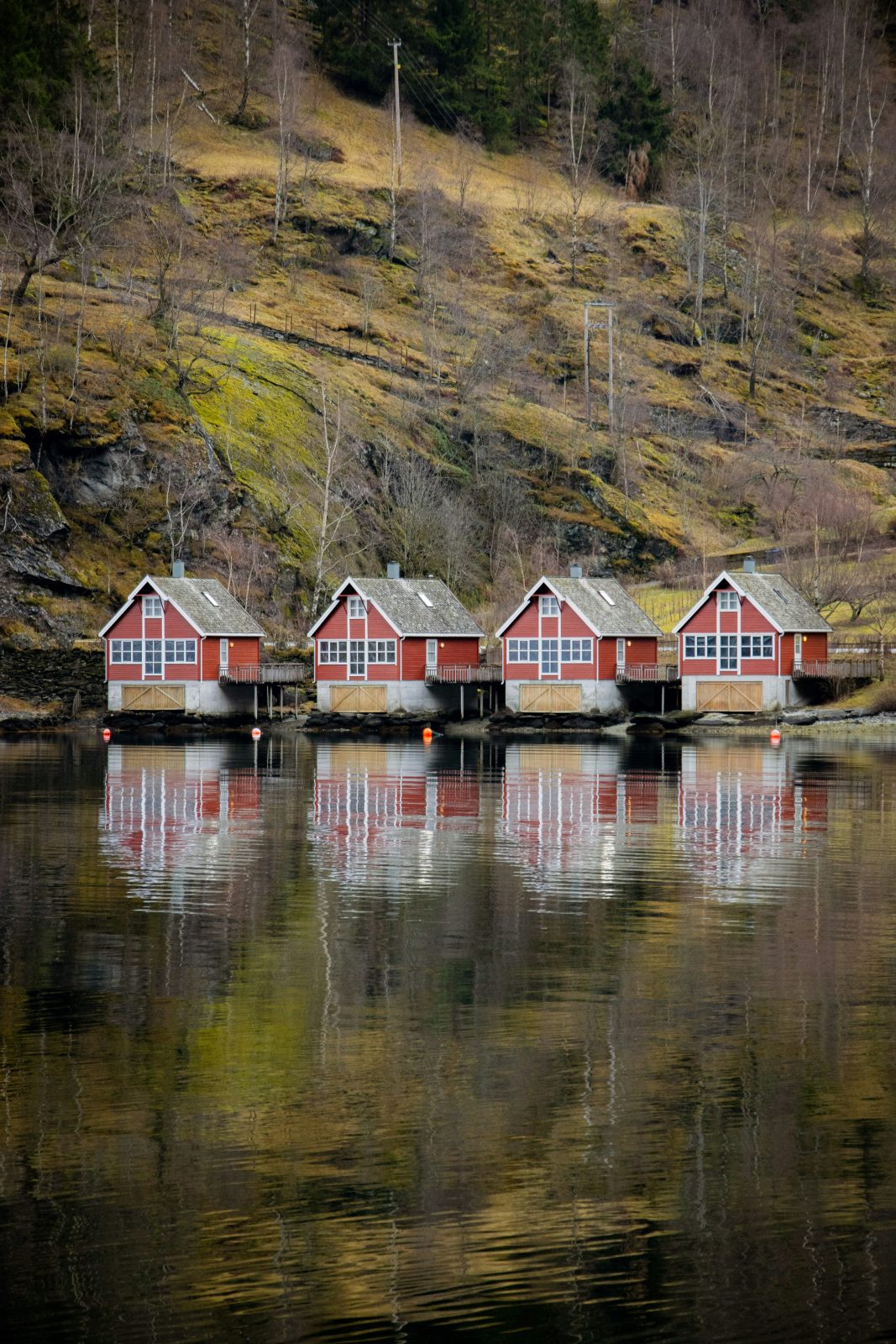 Fire røde hytter på rekke ved vannkanten i Flåm, speilet i det blanke fjordvannet