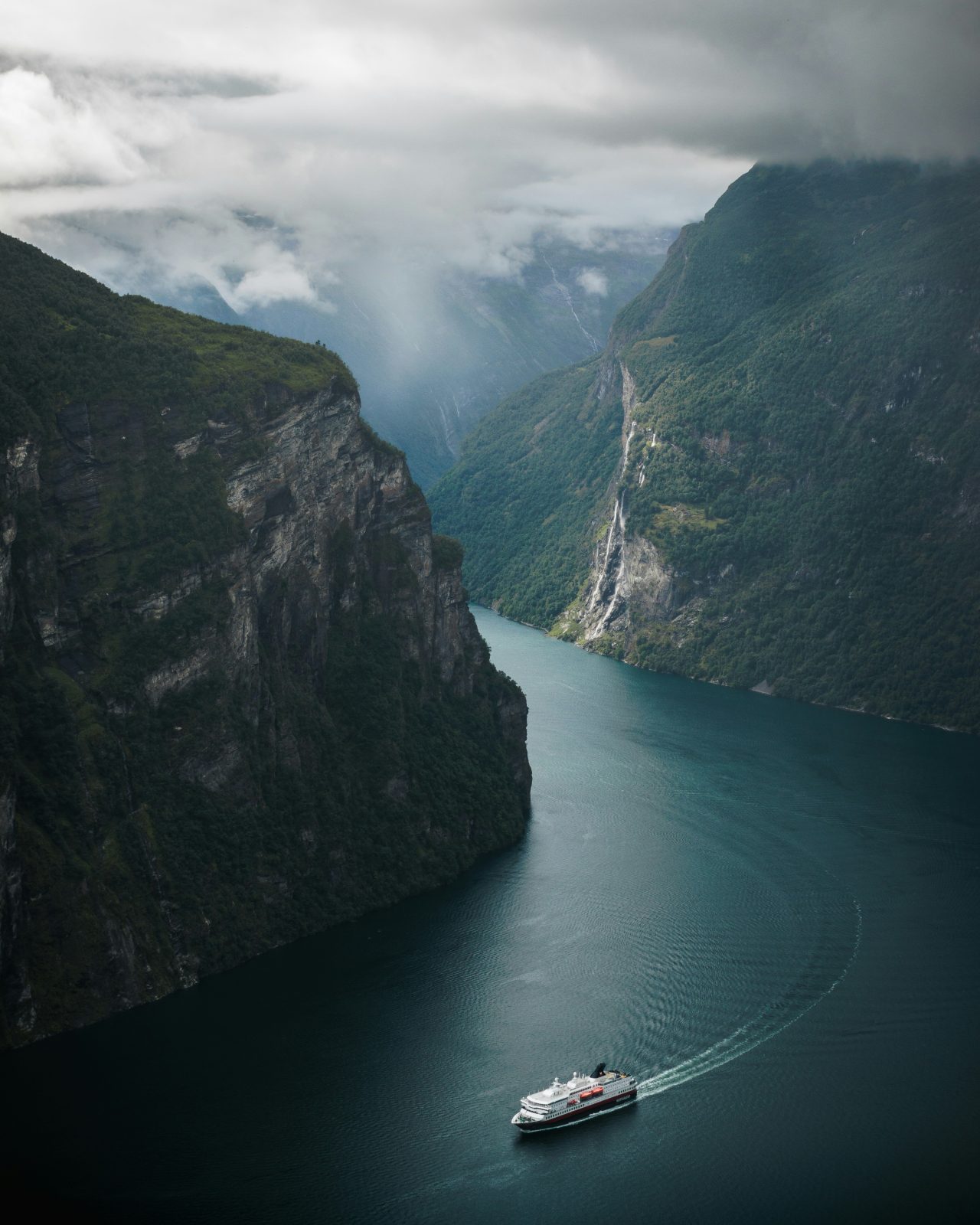 Hurtigruteskip på Geirangerfjorden under lave skyer, med fossefallet De syv søstre i fjellsiden