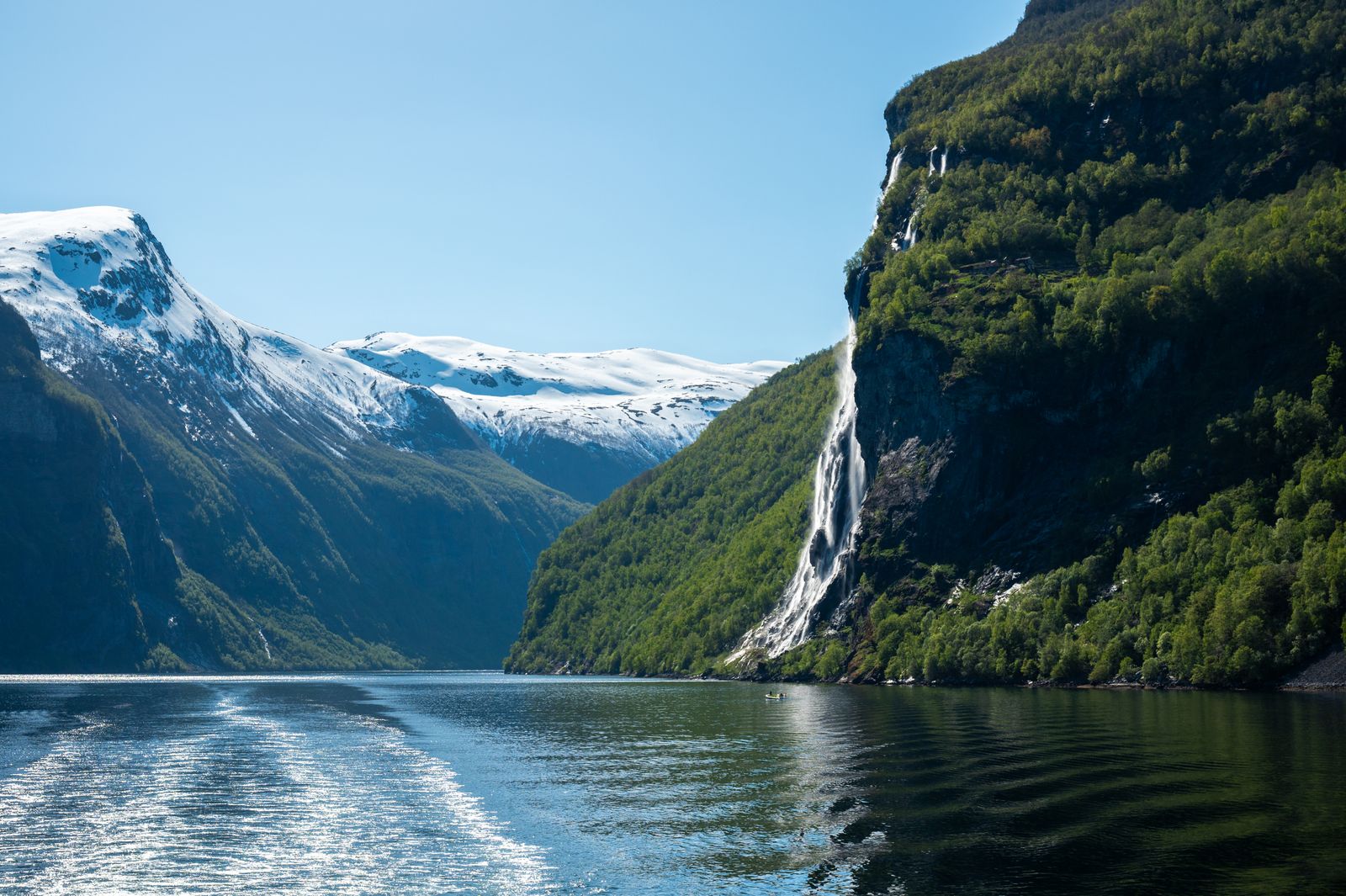 Foss stuper i Geirangerfjorden med snødekte fjell bak