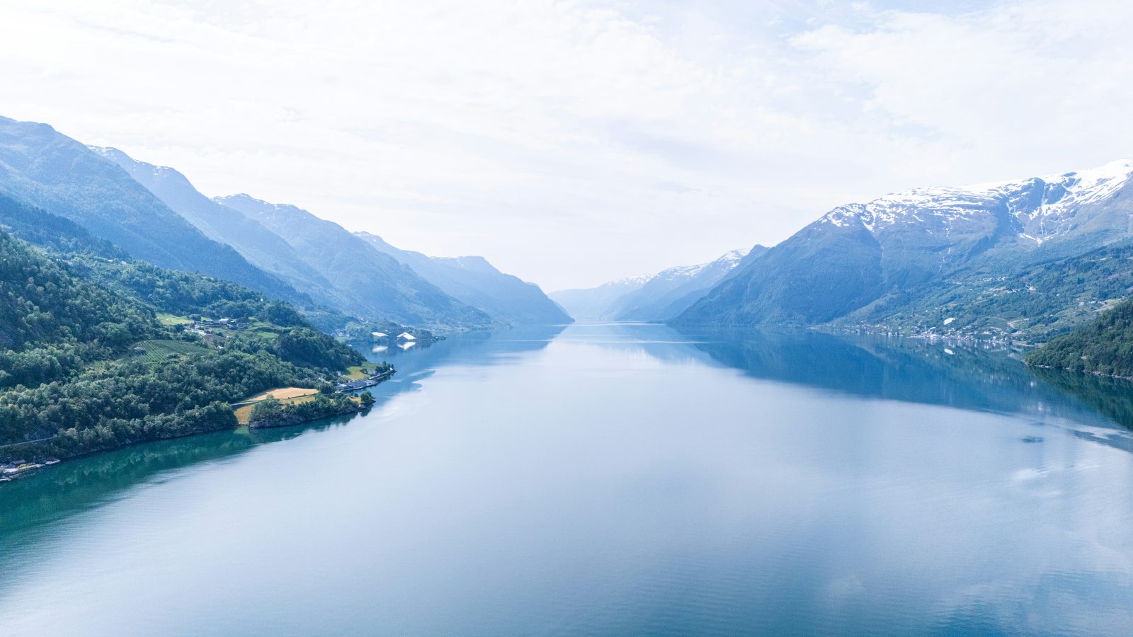 Hardangerfjorden strekker seg innover i sommerdisen, med frukthager i liene og snøkledde fjell på begge sider