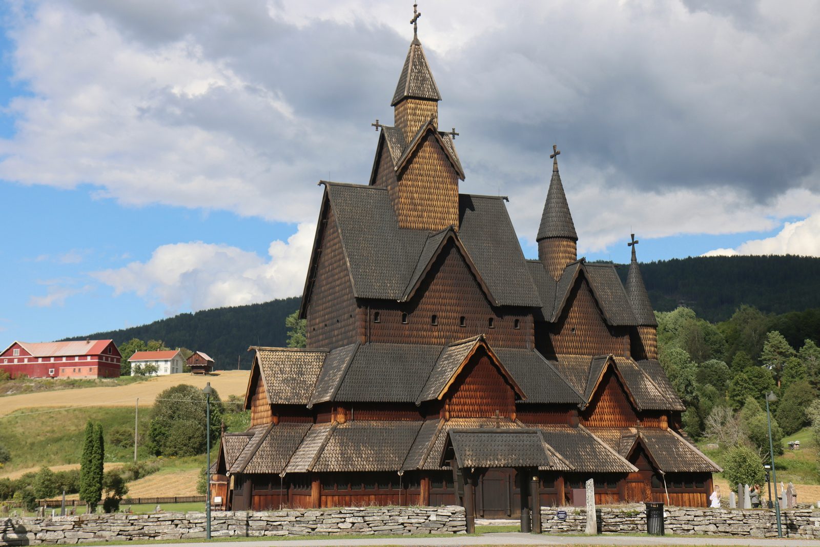 Heddal stavkirke en sommerdag, med takryttere, spir, sponkledde takflater og gårdslandskapet i Telemark bak