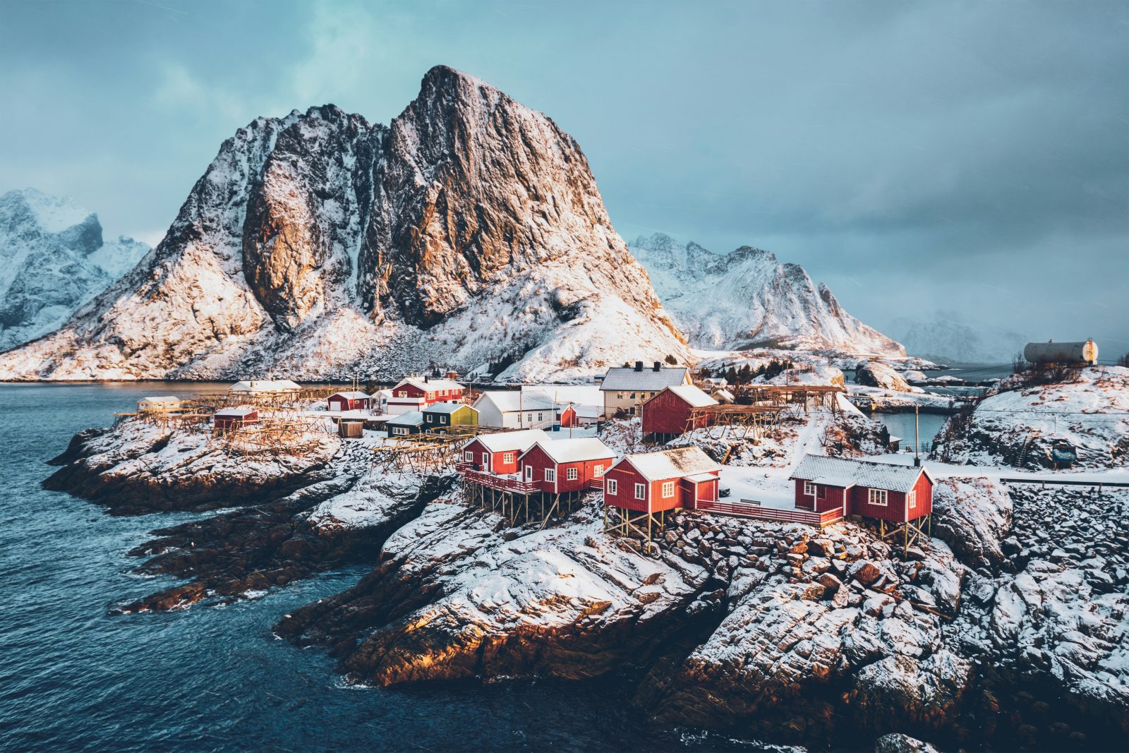 Røde rorbuer på snødekte klipper ved Hamnøy, med tørrfiskhjeller og bratte vinterfjell bak