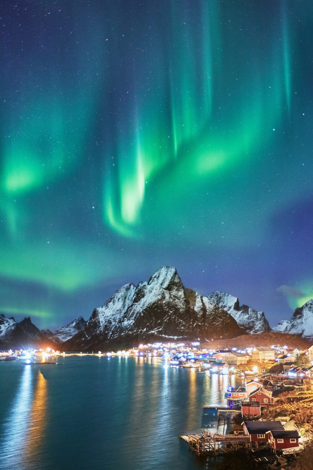 Grønt nordlys danser over de snødekte tindene og de opplyste husene i Reine