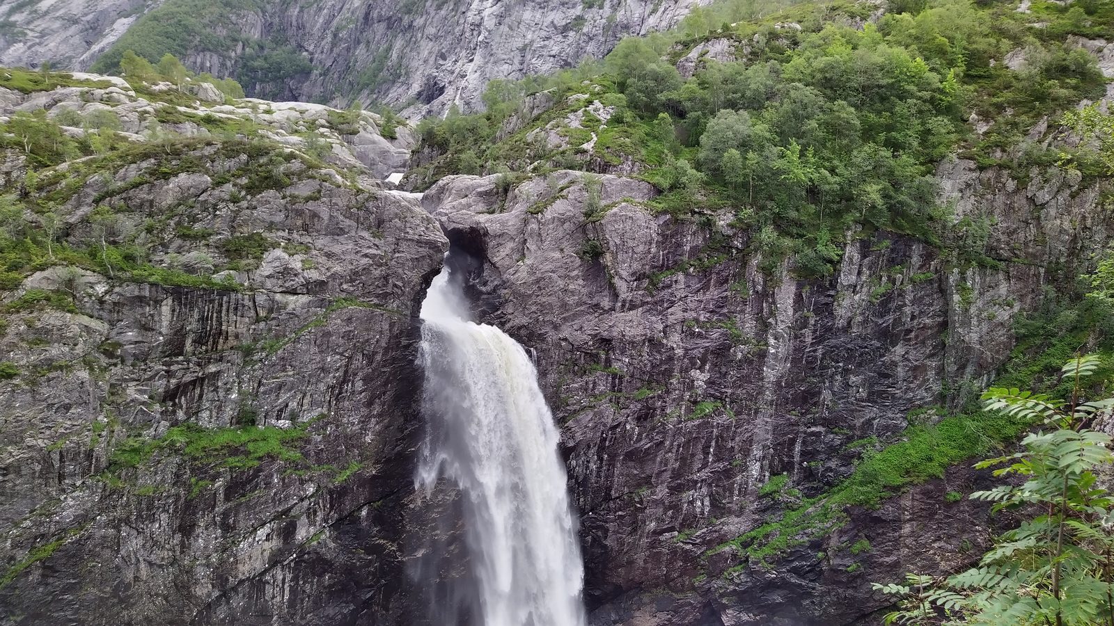 Vannet bryter ut av en revne i berget og faller ned mot juvet, med skogkledde lier og fjell over