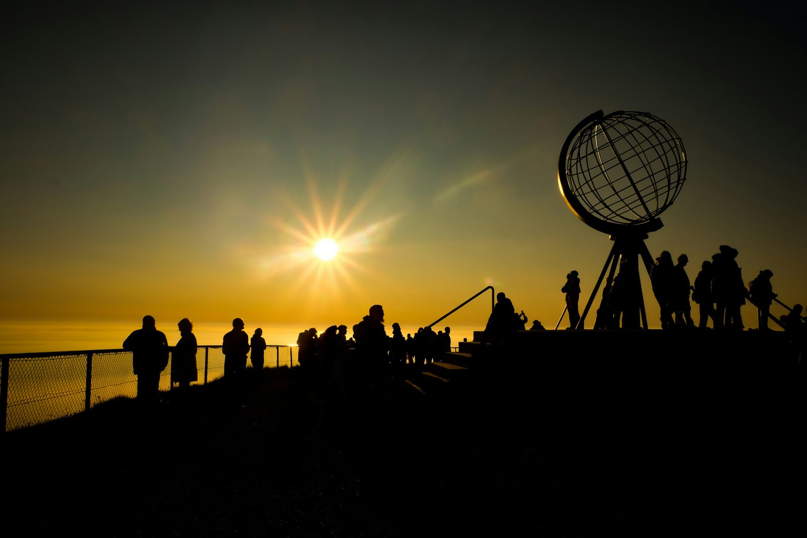 Folkemengde i silhuett rundt Globen på Nordkapp, med midnattssola lavt over havet