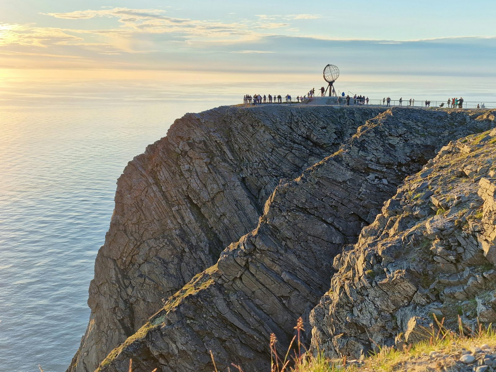 Nordkapplatået med Globen ytterst på klippekanten i kveldssol, med besøkende langs gjerdet og havet bak
