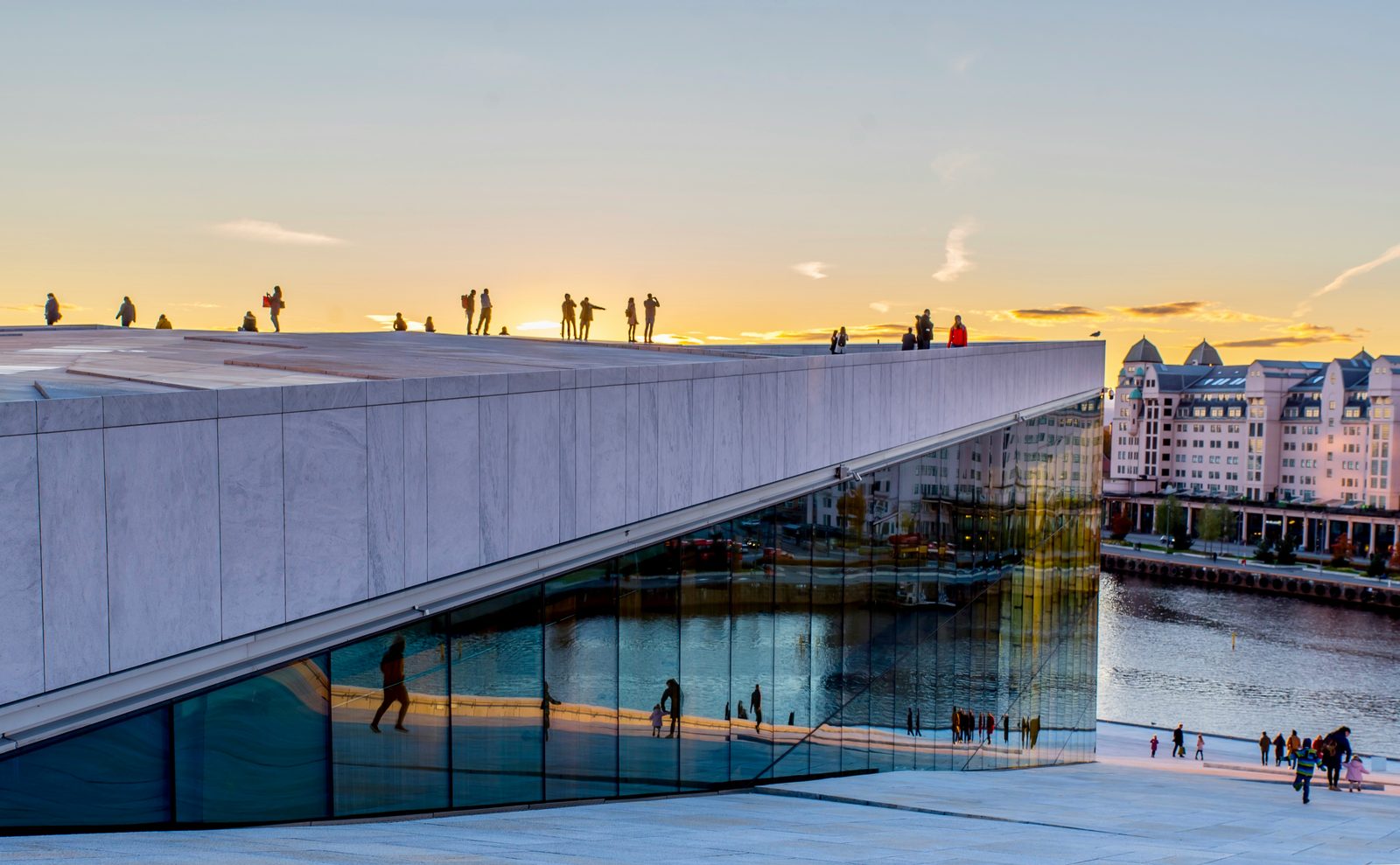 Folk går på Operahusets skrånende marmortak i solnedgang, med byen speilet i glassfasaden