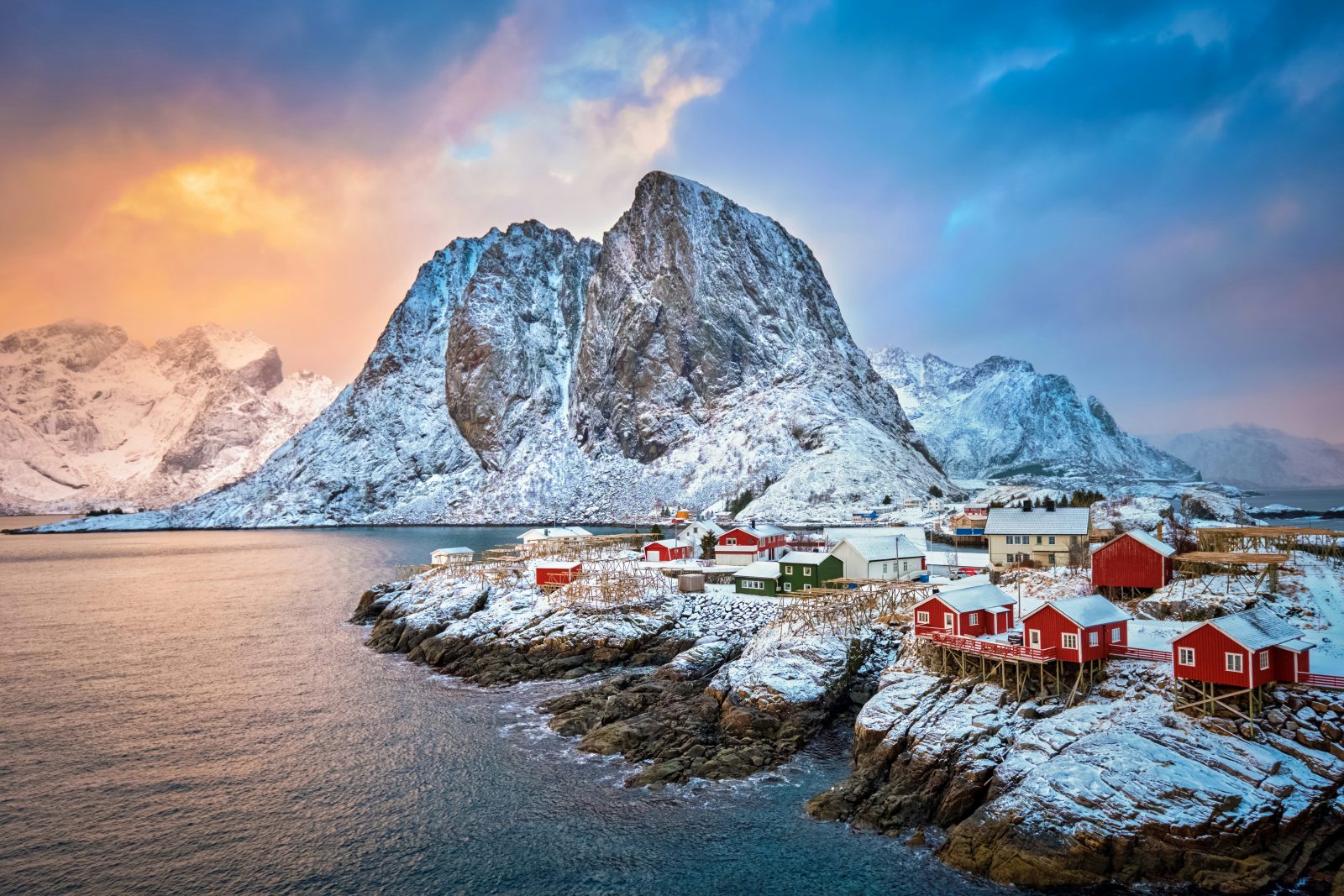 Røde rorbuer på Hamnøy under det bratte fjellet Festhelltinden, i lav vintersol med dramatisk himmel