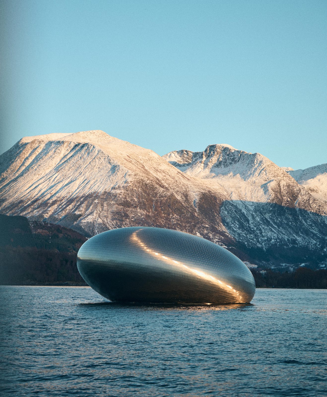 Salmon Eye flyter i Hardangerfjorden med snøkledde fjell bak, stålfasaden lyser i kveldssola