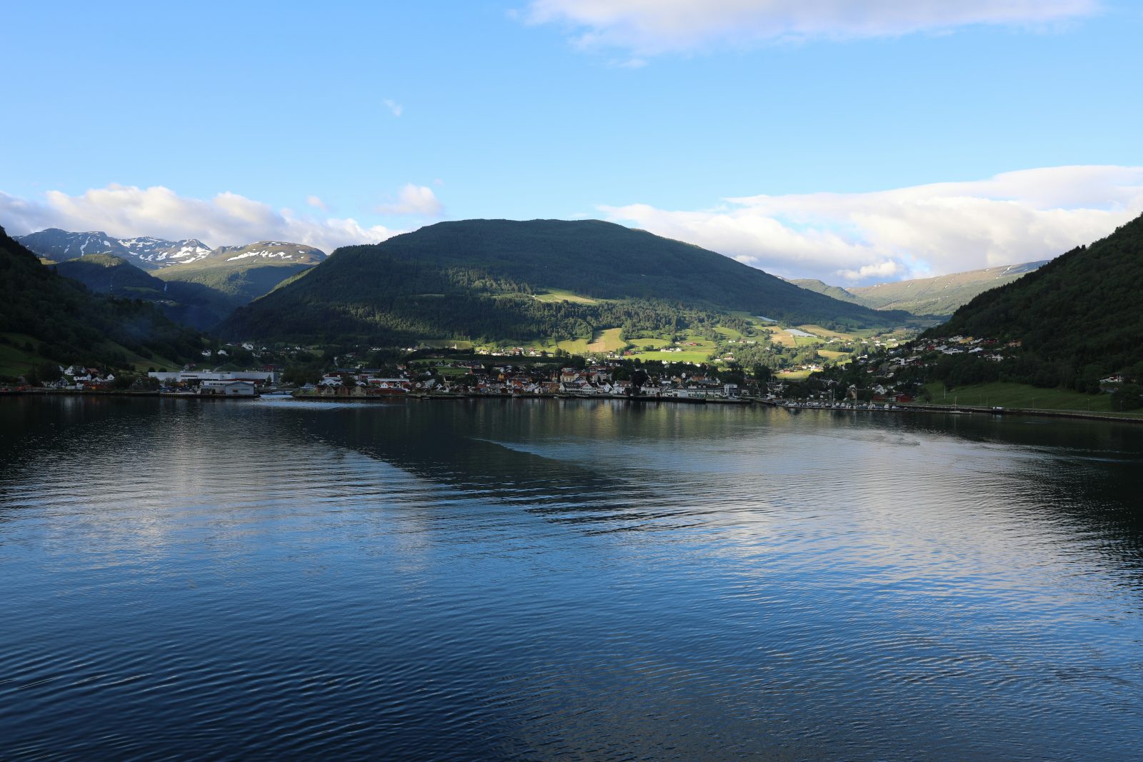 Ei fjordbygd med hvite hus langs vannkanten i kveldslys, med grønne lier og snøkledde fjell bak