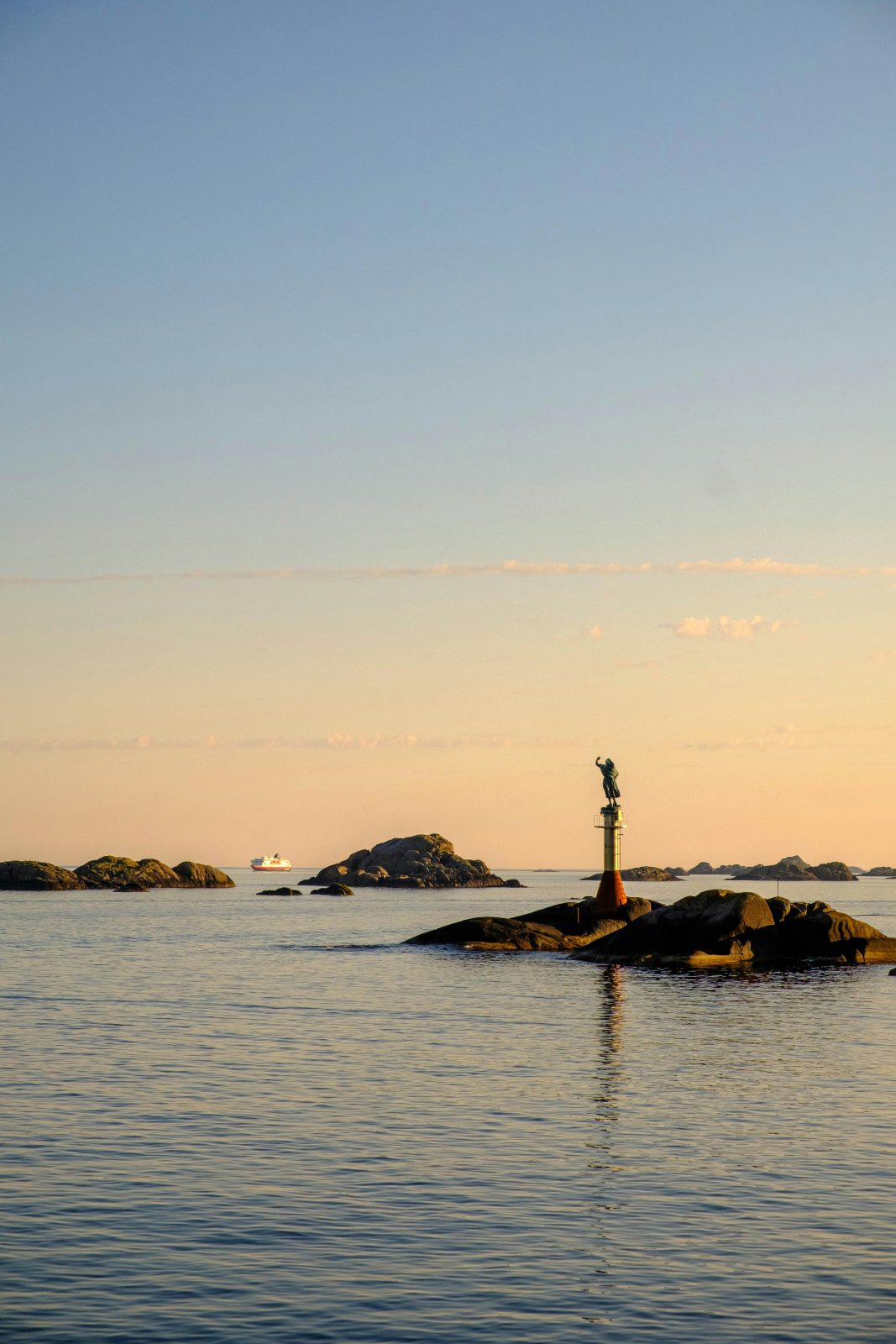 Statuen Fiskarkona på skjæret ved havneinnløpet i Svolvær i midnattssol, med et kystruteskip i horisonten
