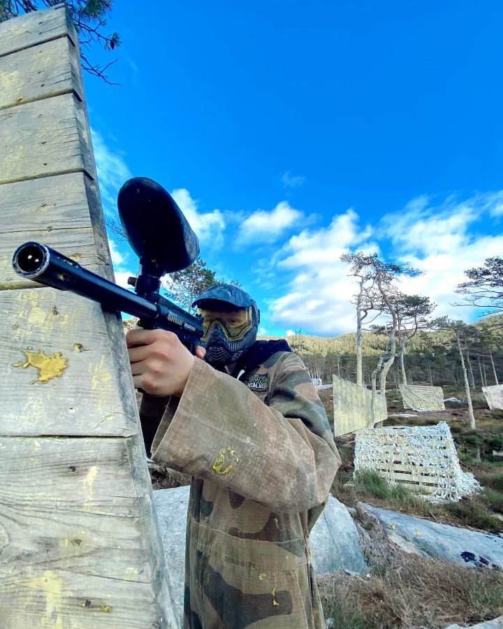 Paintballspiller med maske og markør sikter fra dekning bak en trevegg