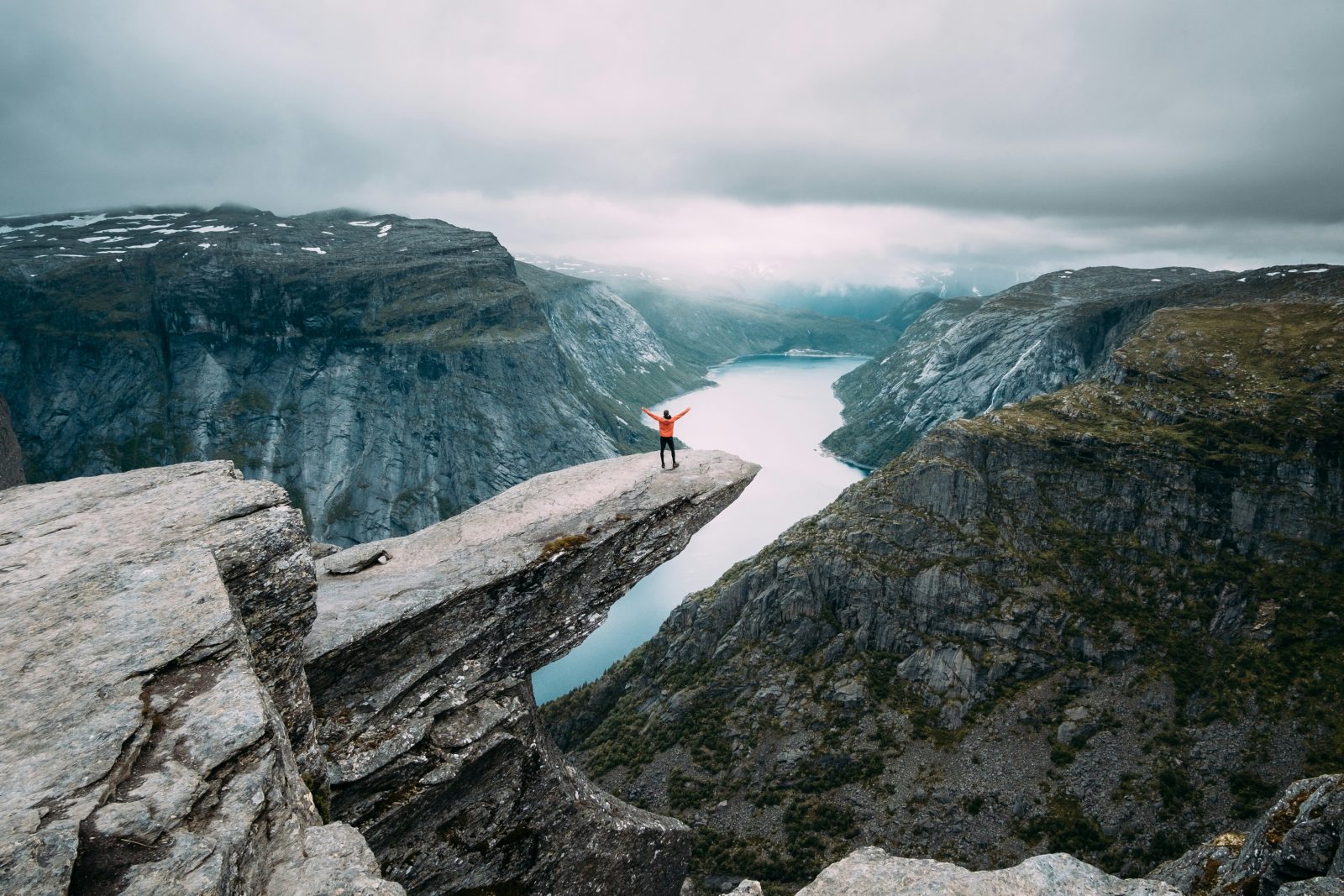 Turgåer i oransje jakke med armene hevet ytterst på Trolltunga under tunge, grå skyer