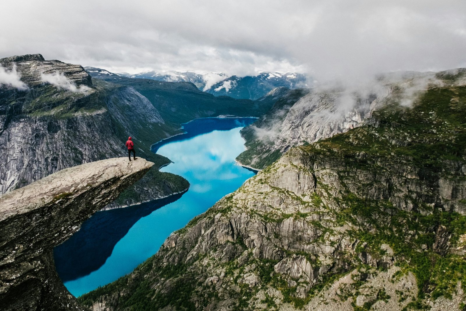 Turgåer i rød jakke står ytterst på Trolltunga med det turkise Ringedalsvatnet dypt der nede