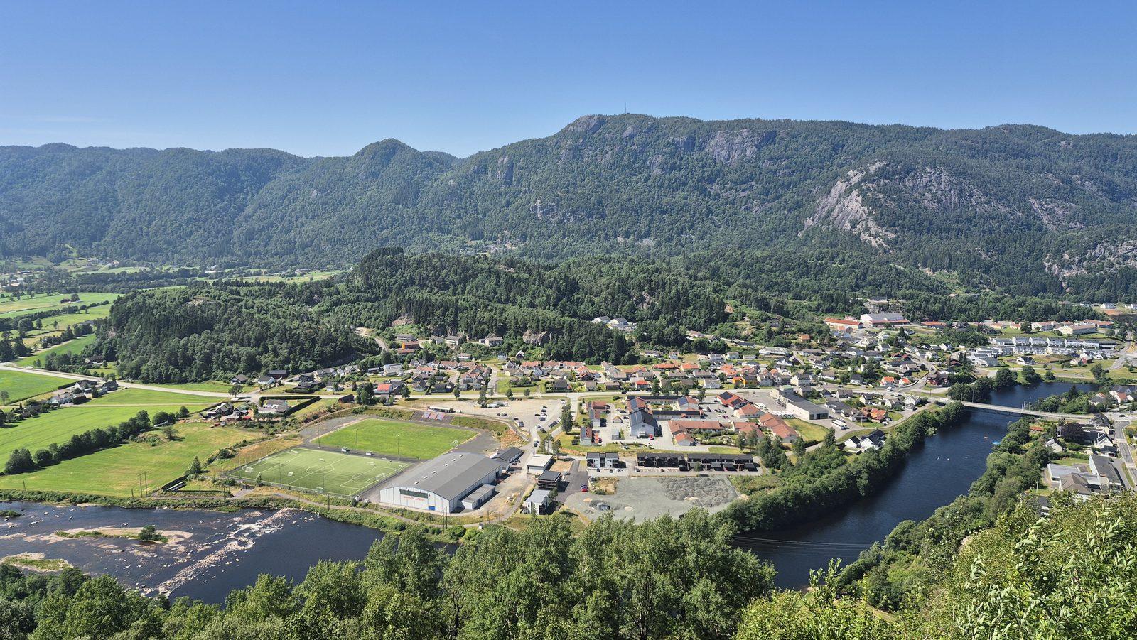 Utsikt fra Utsikten hotell innover Kvinesdalen mot Liknes sentrum, med idrettsanlegg, boligfelt og skogkledde fjell